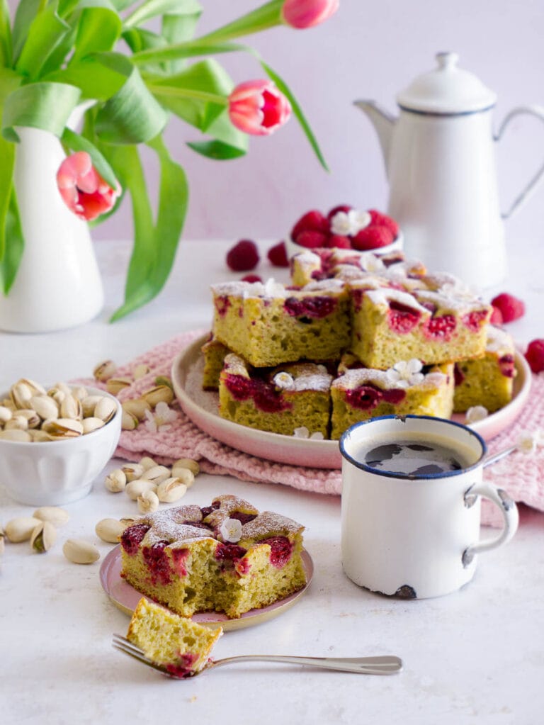 Protein Pistazienkuchen mit Himbeeren in Stückchen geschnitten auf einem Teller, davor liegt ein einzelnes Stückchen auf einem kleinen Tellerchen das bereits begonnen wurde mit einer Kuchengabel zu essen