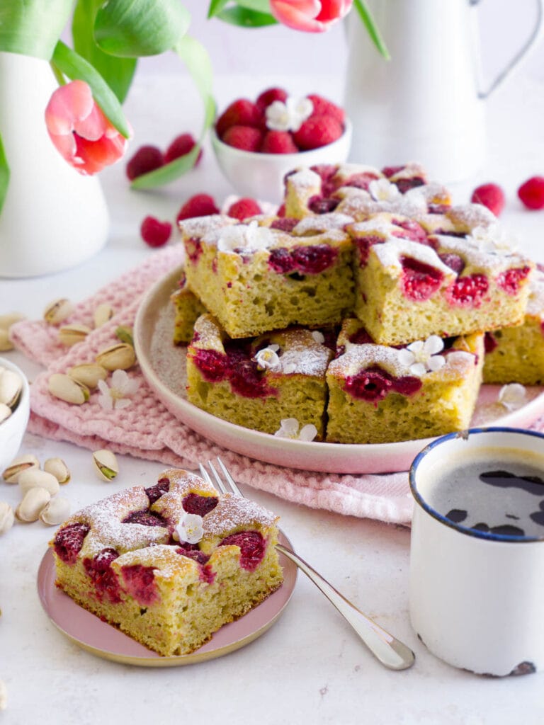 Protein Pistazienkuchen mit Himbeeren in Stückchen geschnitten auf einem Teller, davor liegt ein einzelnes Stückchen auf einem kleinen Tellerchen